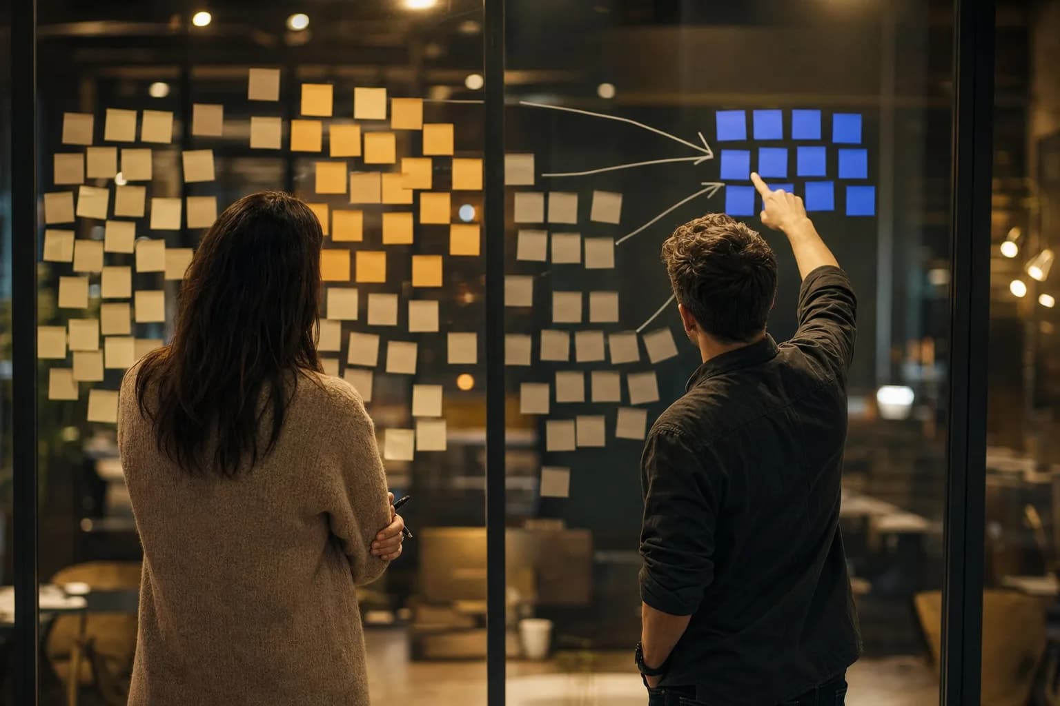 Two colleagues seen from behind at a glass wall covered in sticky notes, one pointing at a single highlighted cluster set apart from competing groups of notes.